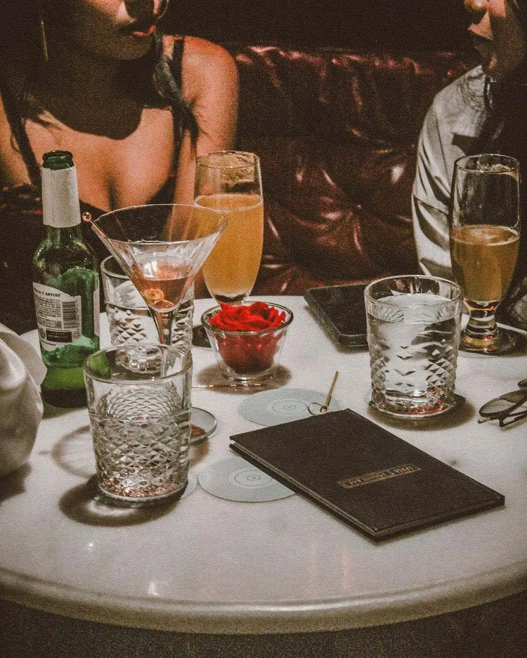 Casual social scene with assorted drinks on a table in a dimly lit bar, two people conversing in the background